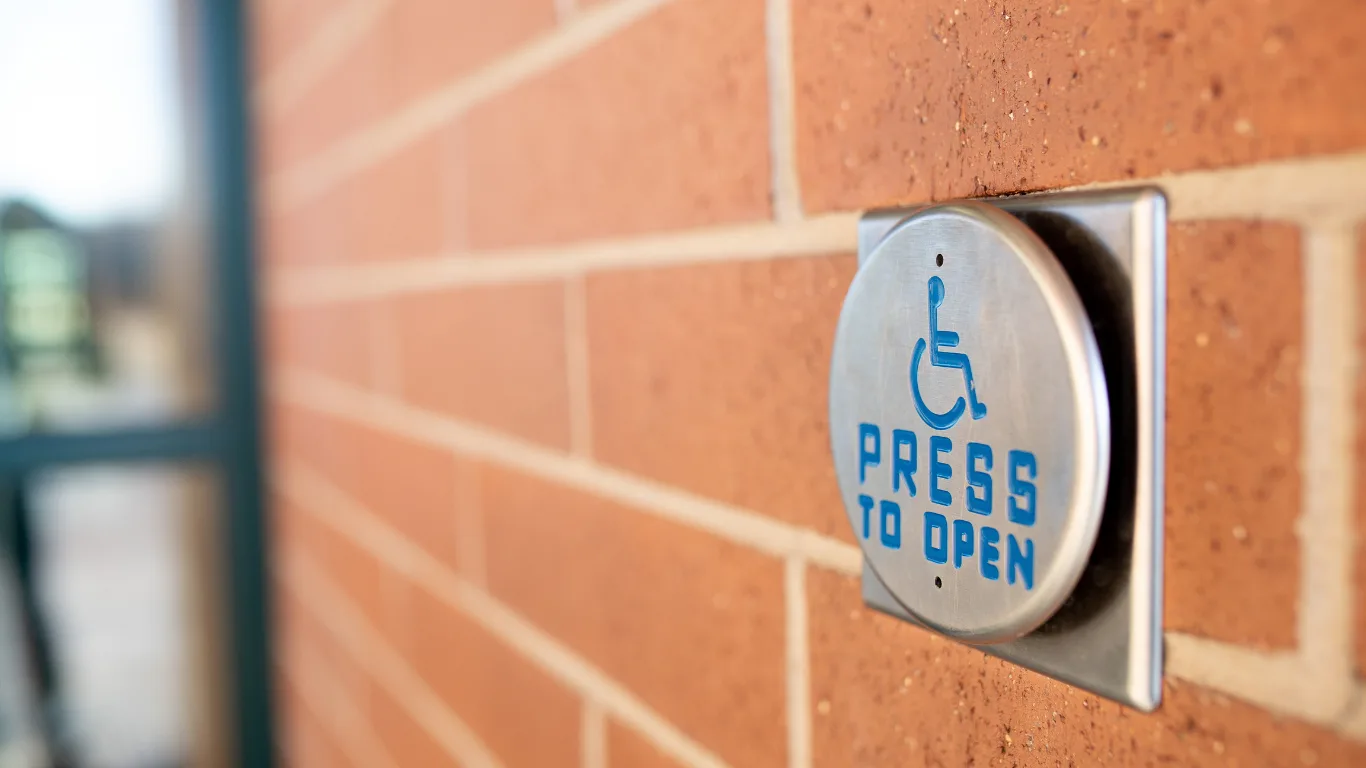 A round metal wheelchair-accessible door button reading &lsquo;Press to Open&rsquo; with the blue wheelchair symbol, mounted on a red brick exterior wall