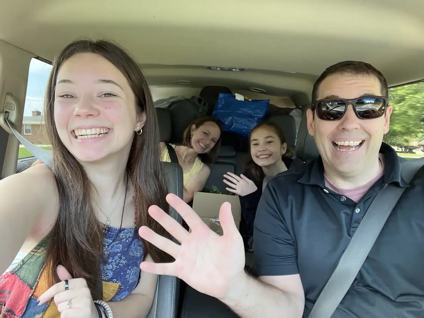 A joyful family of four packed into a car with moving boxes and belongings visible in the back, smiling and waving at the camera on a move-in day drive