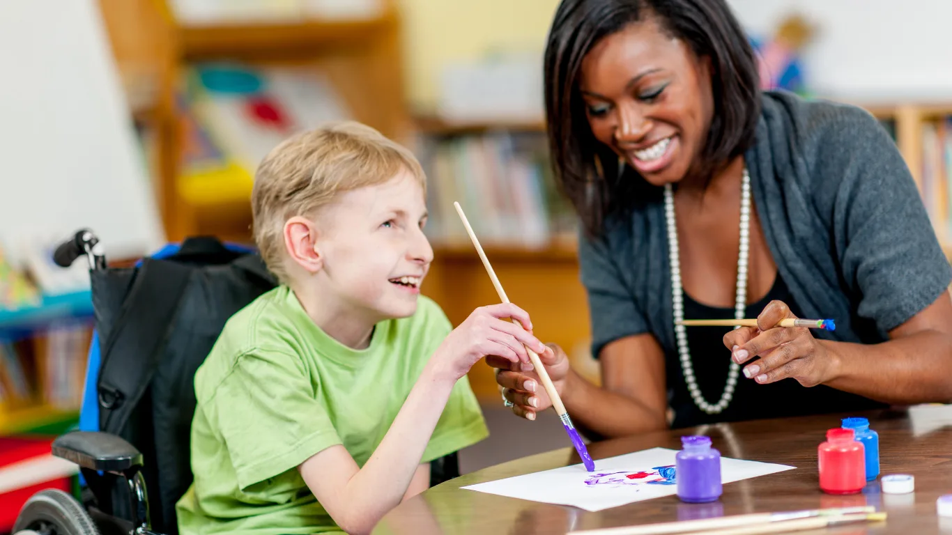 A smiling boy in a green shirt seated in a wheelchair paints on paper with a smiling woman beside him in a colorful classroom lined with bookshelves