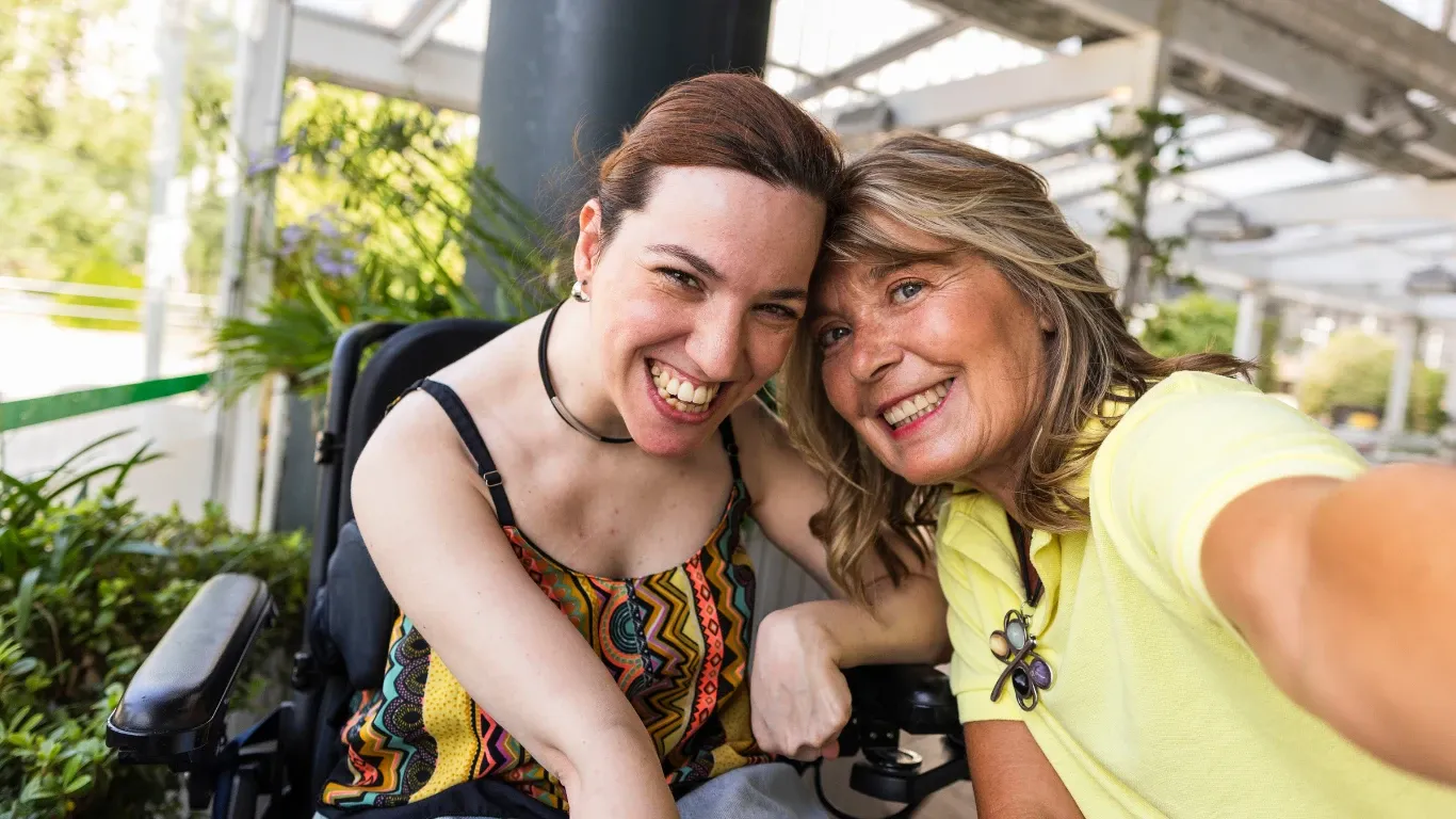 Two women smiling and leaning their heads together for a selfie, one seated in a power wheelchair