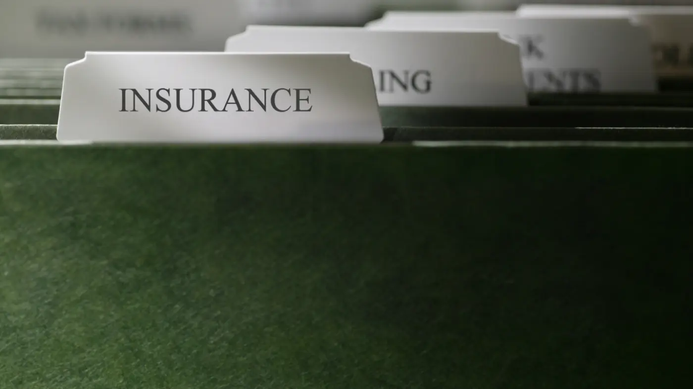 Close-up of a green hanging file folder with a tab labeled &lsquo;INSURANCE&rsquo; visible in the foreground and other file tabs blurred behind it