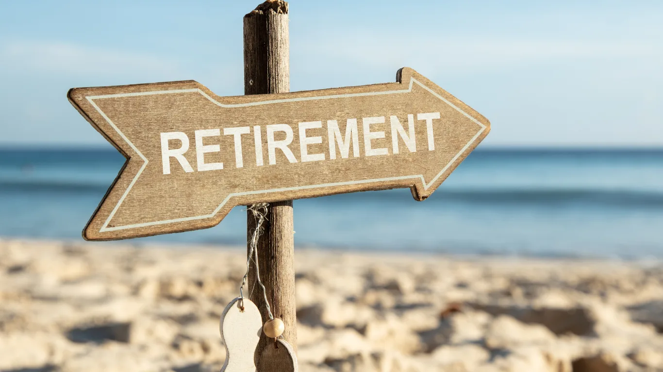 A wooden directional sign with &lsquo;Retirement&rsquo; painted on it, planted in the sand on a beach with the ocean and clear sky in the background