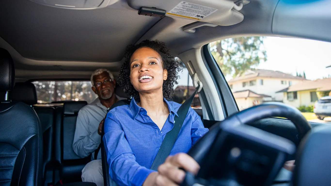 A smiling woman in a blue shirt drives a car with an older man seated in the back, on a sunlit suburban street