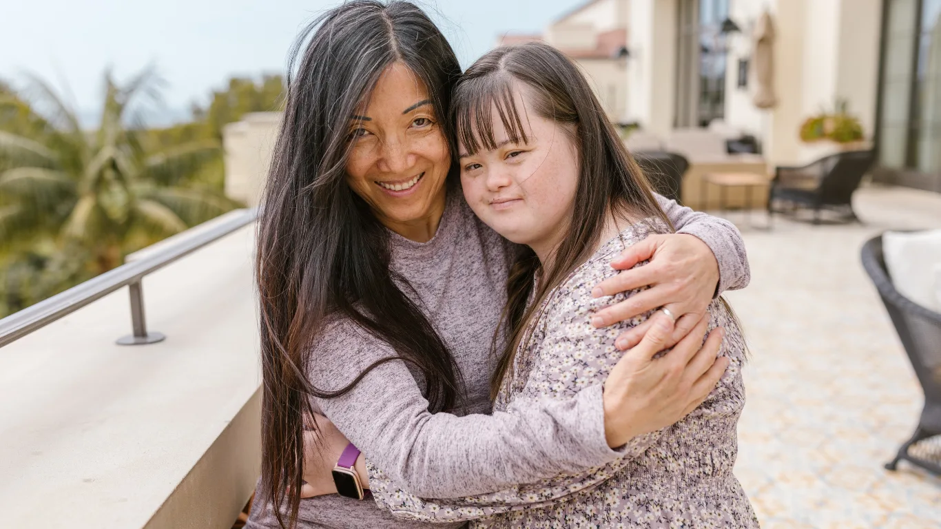 A mother and her adult daughter embracing on a sunlit balcony
