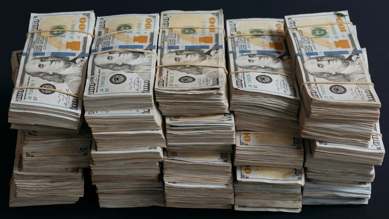 Several large stacks of $100 bills bundled with rubber bands, arranged on a dark surface